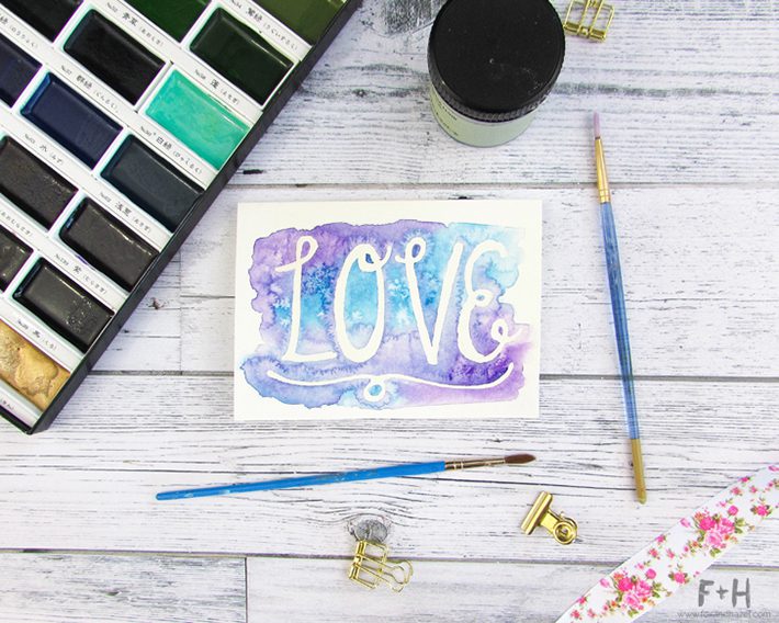 watercolor valentine card with the word LOVE on white wood background surrounded by art supplies