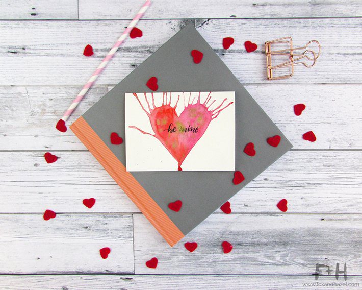 watercolor heart valentine card on top of grey book on white wood background