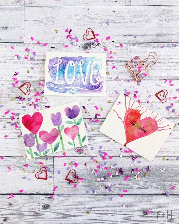 three watercolor valentines cards surrounded by confetti on white wood background