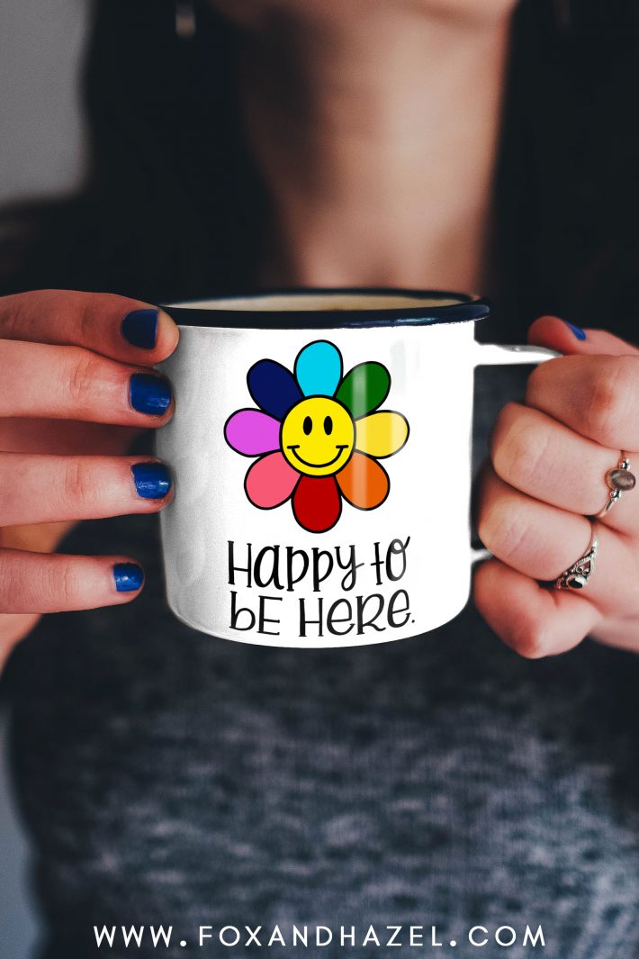 woman holding a smiley flower coffee mug with words "happy to be here" 