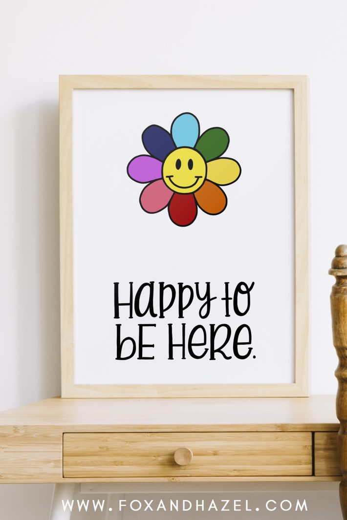 free smiley flower art print in wood frame with words "happy to be here" sitting on a wood desk.