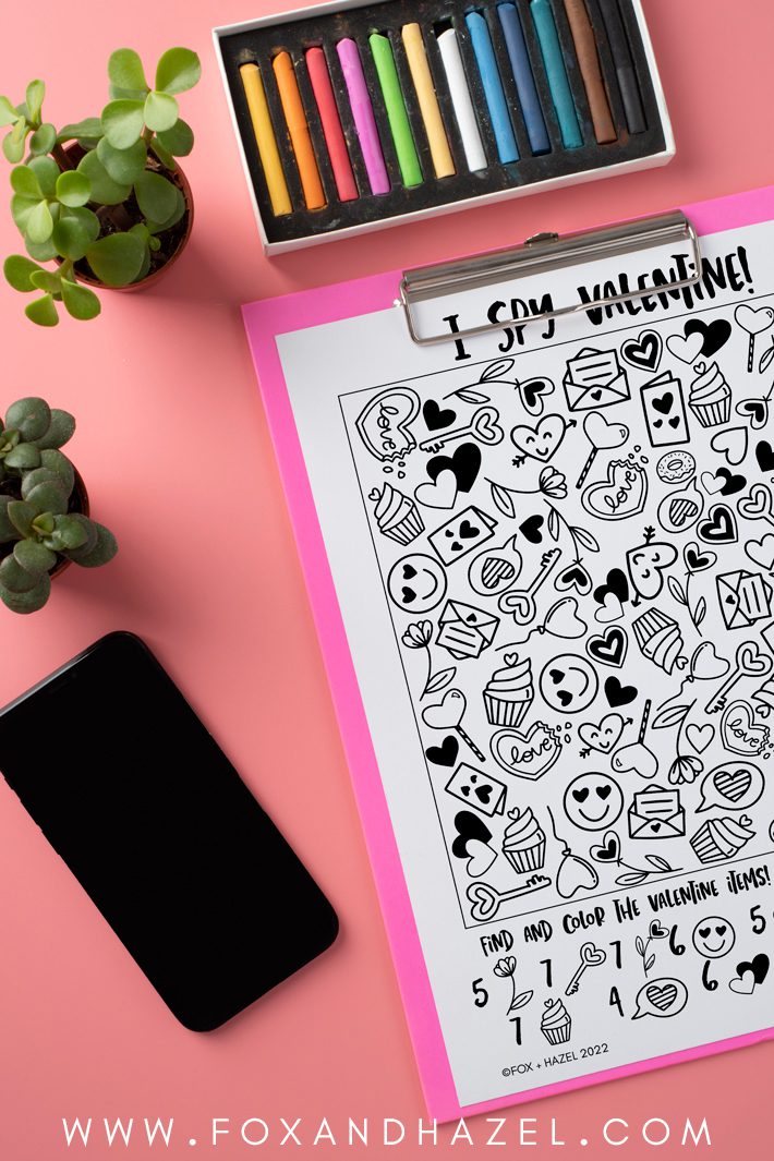 free printable i spy valentine activity sheet on a pink clipboard, laying on a pink background accompanied by a smart phone, small plants and a tray of pastels