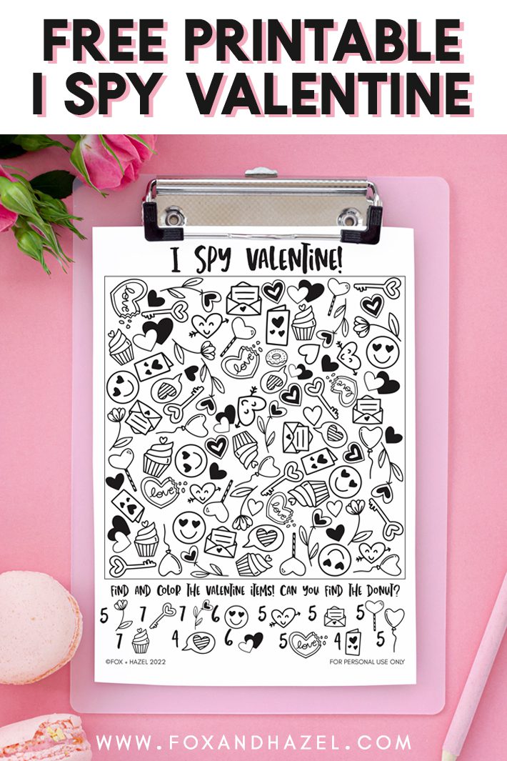free printable valentine I spy activity sheet on a pink clipboard, laying on a pink background surrounded by pink roses and pink macarons